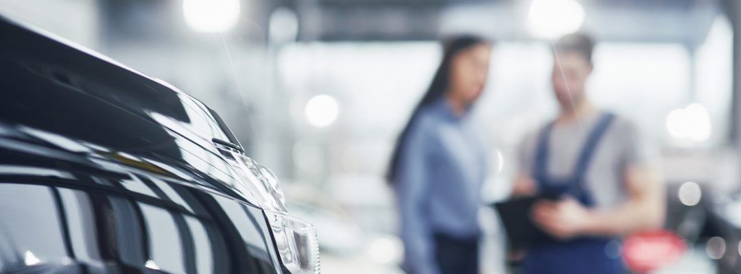 Beautiful young woman is talking to handsome car mechanic while repair a car in dealership.